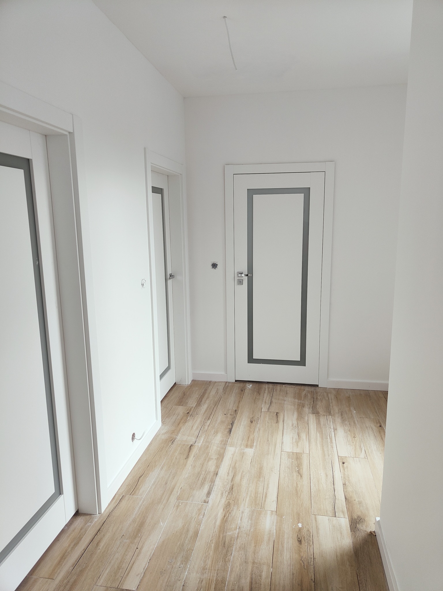 Korytarz w odcieniach bieli z trzema białymi drzwiami z szarymi ramkami i podłogą imitującą drewno, widoczne gniazdko elektryczne i wystający kabel ze ściany