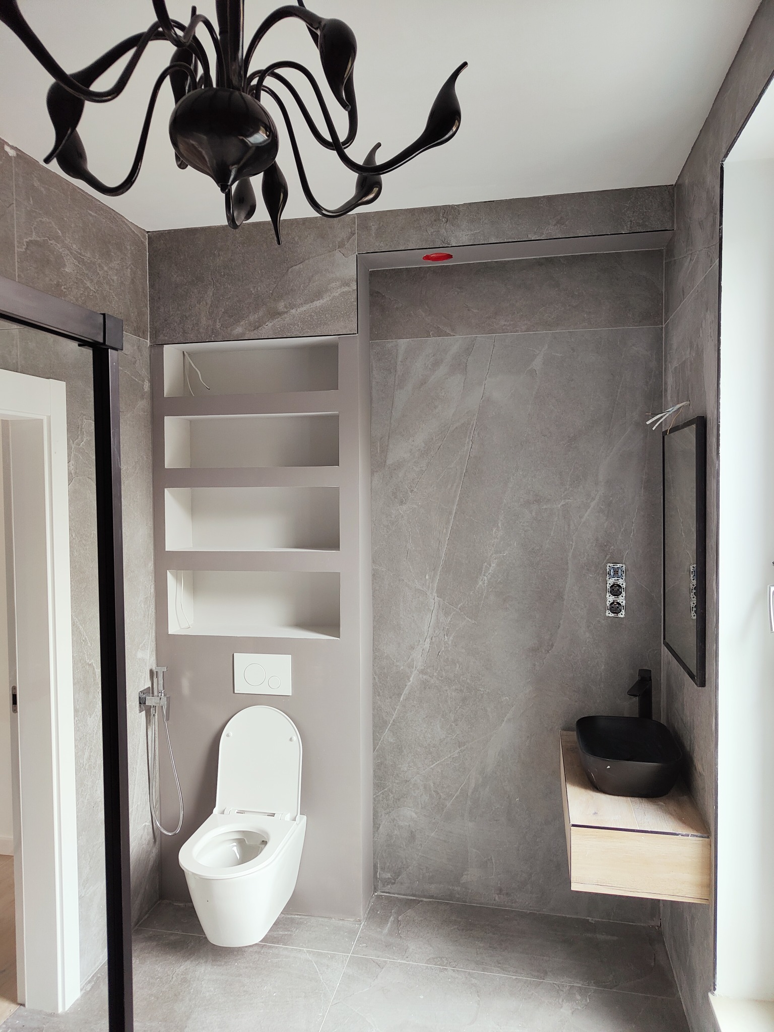 Nowoczesna łazienka wykończona szarym gresem imitującym marmur, z czarną armaturą, podwieszaną toaletą i designerskim żyrandolem.