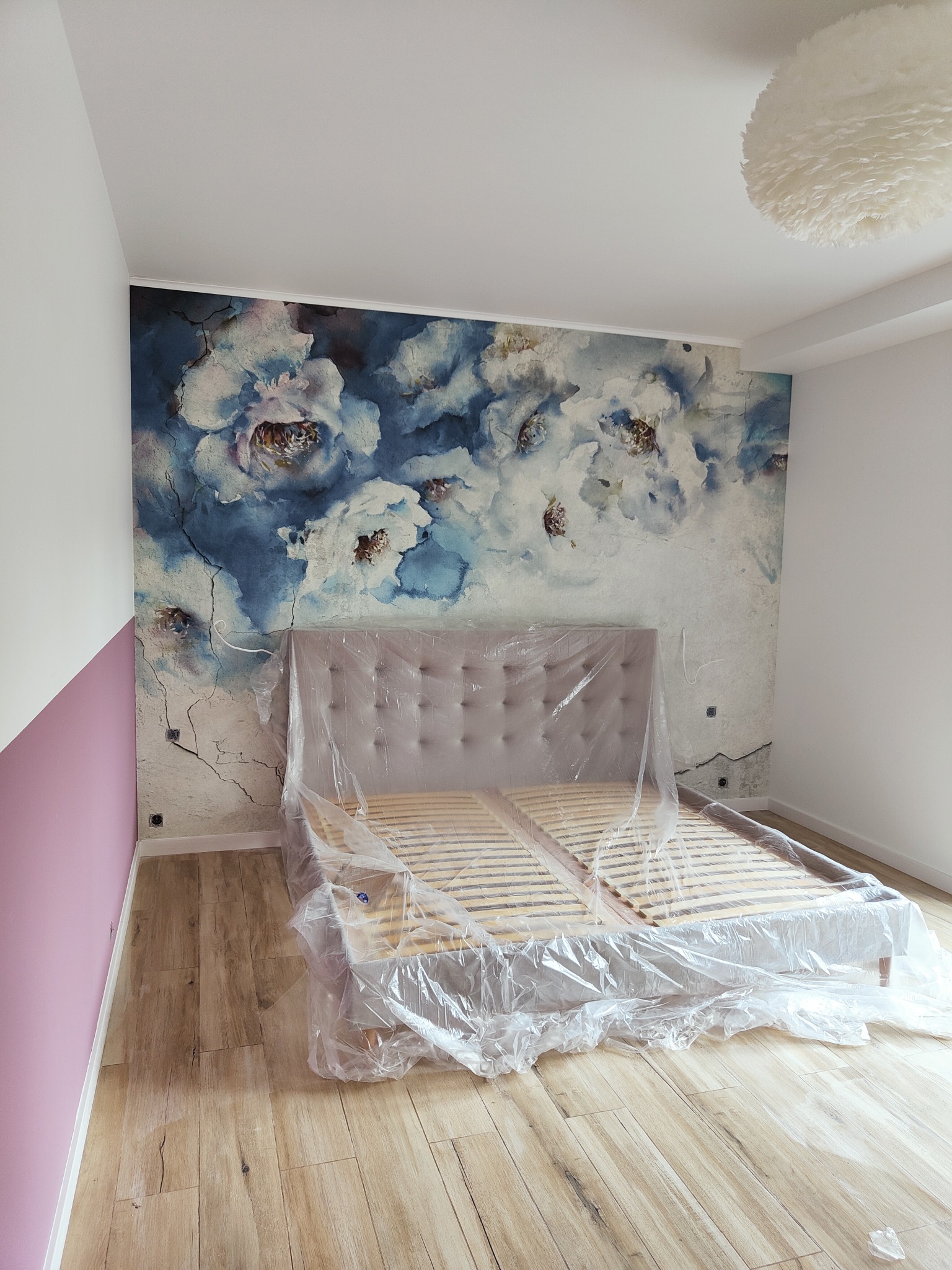 Pokój w trakcie malowania z zabezpieczonym łóżkiem folią, ściana z motywem kwiatowym, fragment ściany pomalowany na różowo.
