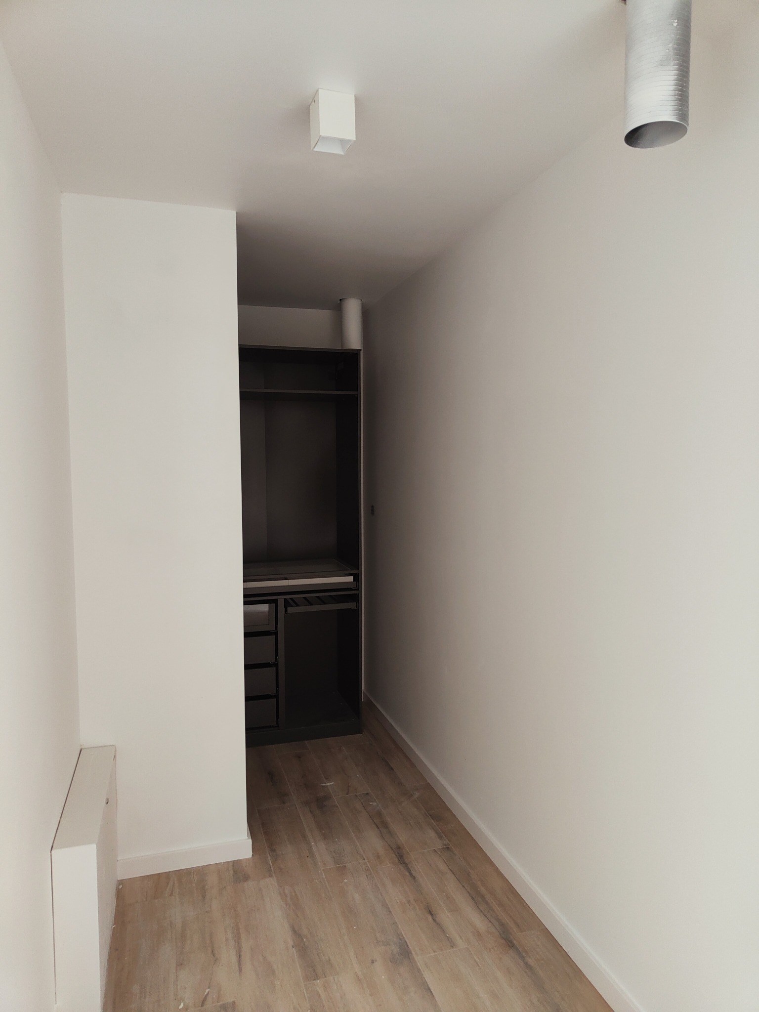 Wnętrze pokoju po remoncie, z pomalowanymi na biało ścianami, drewnopodobną podłogą i wnęką z zabudowaną szafą w ciemnym kolorze. Widoczny biały grzejnik i lampy sufitowe.