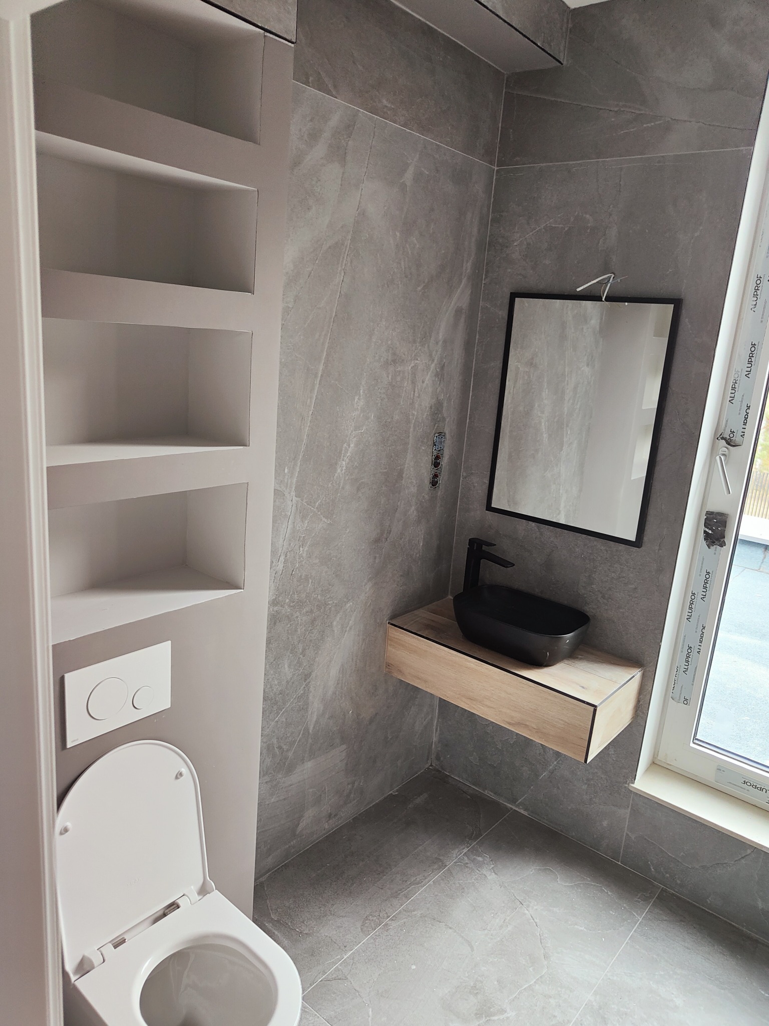 Nowoczesna łazienka z szarą okładziną imitującą marmur, wisząca czarna umywalka na drewnianej szafce, podwieszana toaleta i wnęka z półkami.