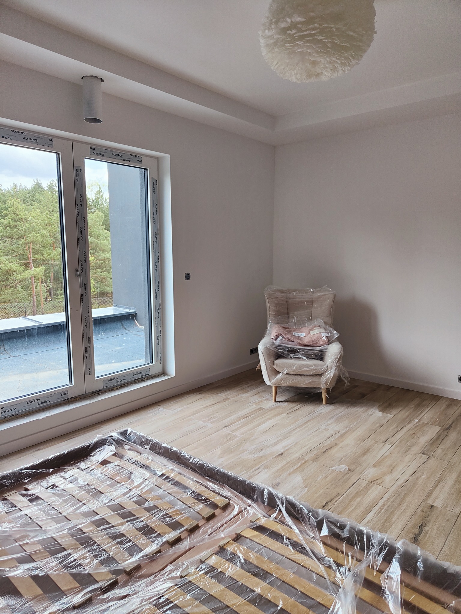 Pokój w trakcie wykańczania: świeżo pomalowane białe ściany, podłoga imitująca drewno, okno z widokiem na las, meble zabezpieczone folią malarską.