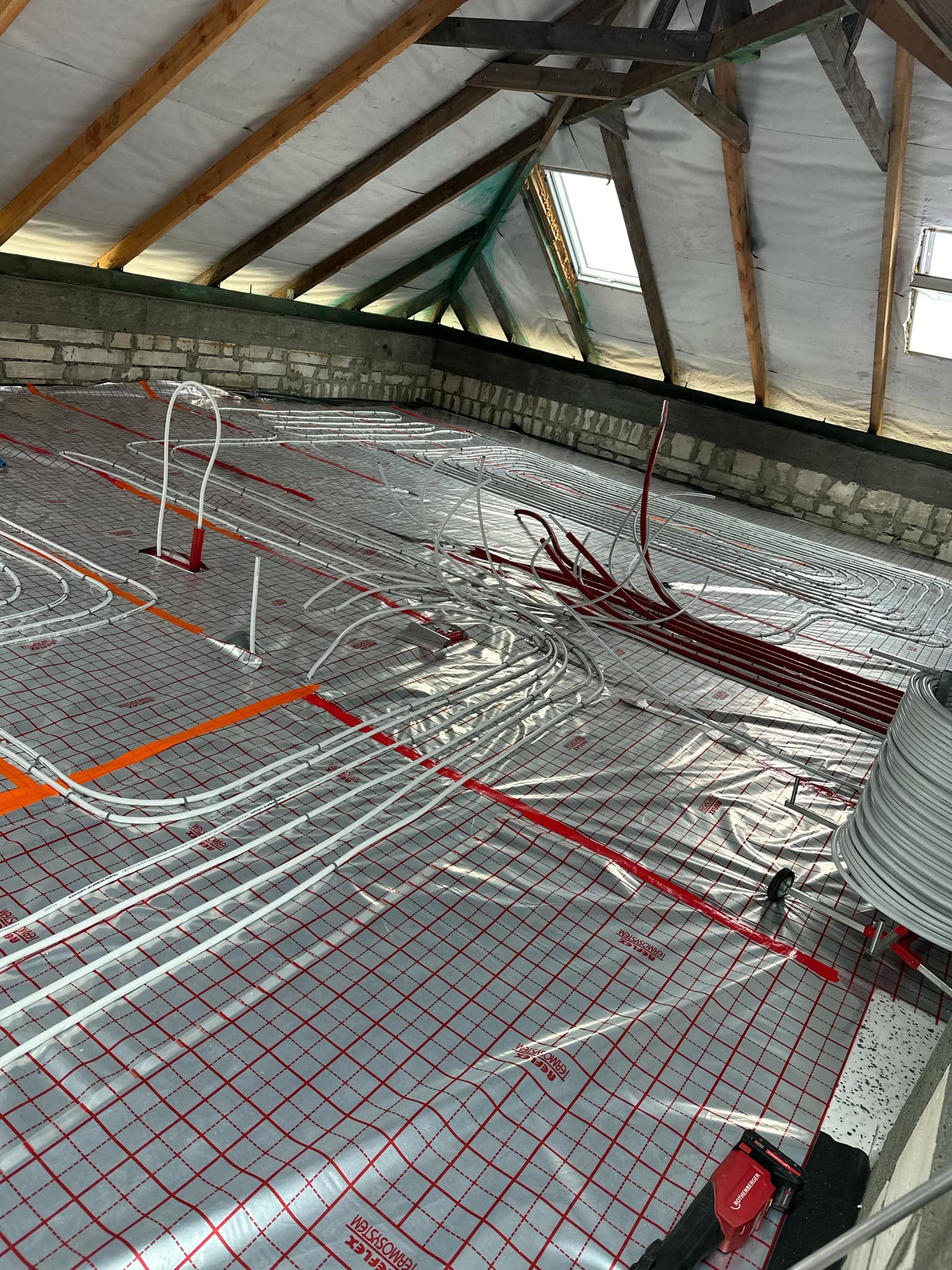 Montaż ogrzewania podłogowego w poddaszu: białe i czerwone rurki ułożone na folii z czerwoną siatką, widoczne krokwie i okna dachowe.
