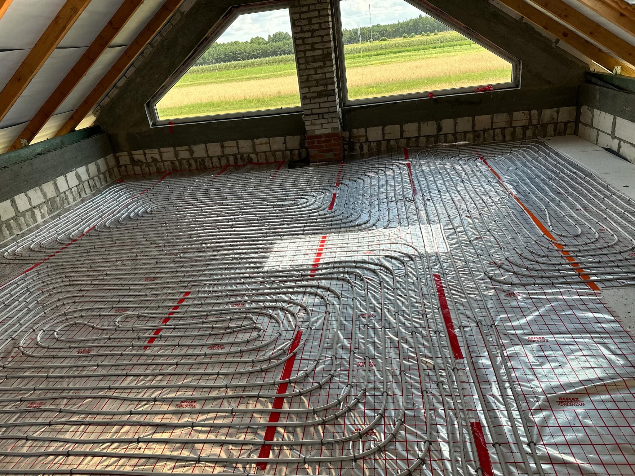 Układanie rur ogrzewania podłogowego na folii z siatką w nowo budowanym poddaszu z widokiem na pole przez okno.