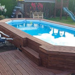 Meble Bertus - Prostokątny basen naziemny z drewnianą obudową i schodkami, z widoczną pompą filtrującą oraz drewnianym tarasem obok, w tle trawnik, huśtawka i zjeżdżalnia.