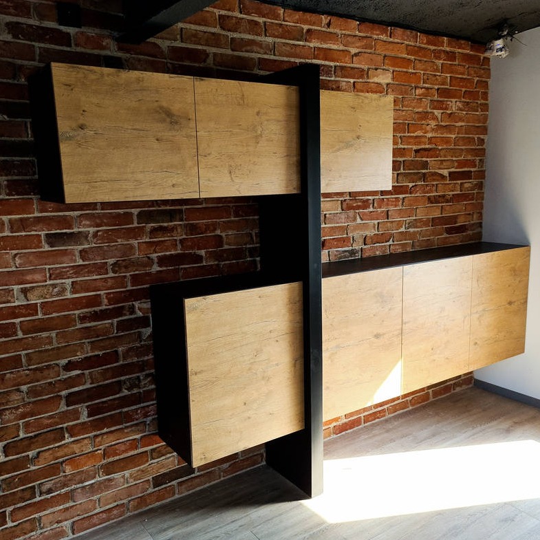 Nowoczesna zabudowa meblowa z frontami w odcieniach jasnego drewna, czarne elementy konstrukcyjne, na tle ściany z czerwonej cegły i jasnej podłogi.