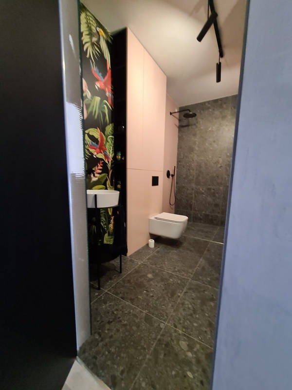 Nowoczesna łazienka z szafką w kolorze pudrowego różu, toaletą podwieszaną i umywalką w szafce z motywem tropikalnym, oświetlenie punktowe na suficie, szare płytki podłogowe i ścienne.