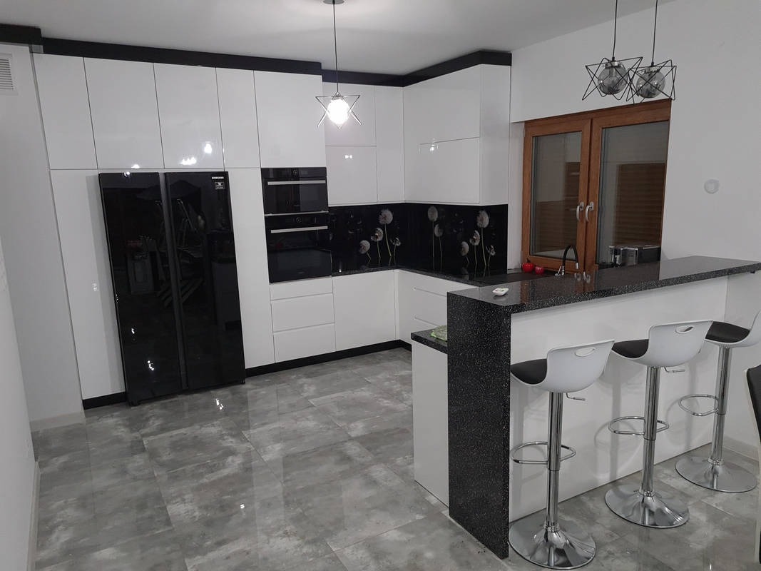 Nowoczesna kuchnia z białymi szafkami, czarnym blatem i panelem dekoracyjnym z motywem dmuchawców, oświetlona wiszącymi lampami w kształcie gwiazd, z barkiem i trzema hokerami.