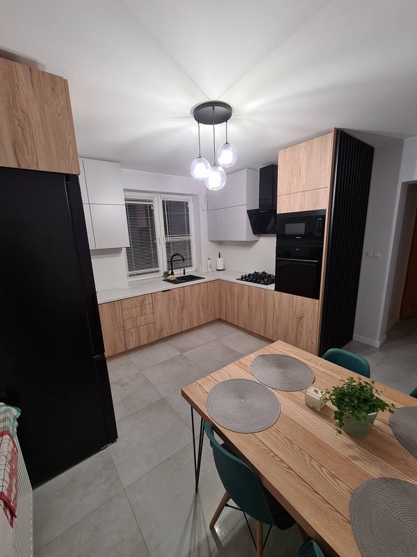 Nowoczesna kuchnia z drewnianymi frontami szafek, białymi szafkami wiszącymi i czarnym sprzętem AGD, oświetlona lampą wiszącą z trzema szklanymi kulami, widok z perspektywy stołu z zielonymi...