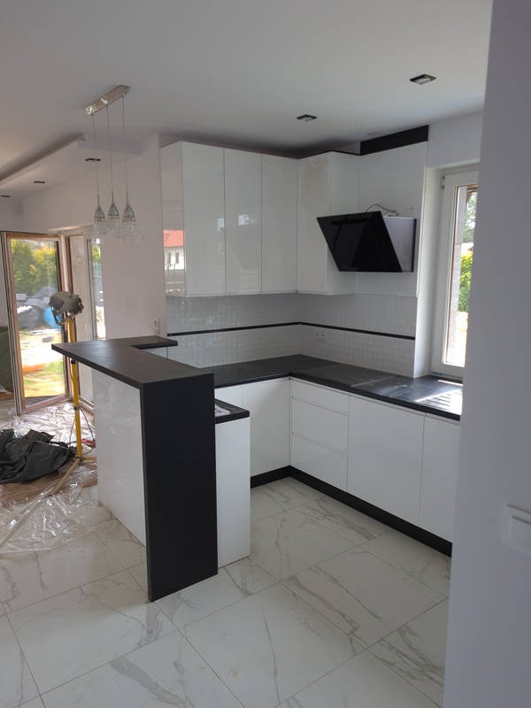 Nowoczesna, biała kuchnia z czarnymi blatami i barkiem, wykończona mozaiką, widok na ogród przez okno, w trakcie montażu.