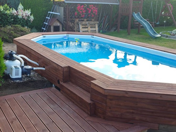 Prostokątny basen naziemny z drewnianą obudową i schodkami, z widoczną pompą filtrującą oraz drewnianym tarasem obok, w tle trawnik, huśtawka i zjeżdżalnia.