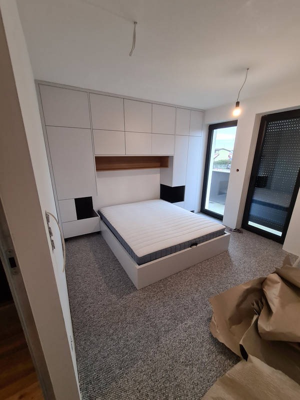 Jasna sypialnia z białą zabudową meblową obejmującą szafę i ramę łóżka z drewnianą wnęką nad wezgłowiem, widok na balkon przez duże okno.