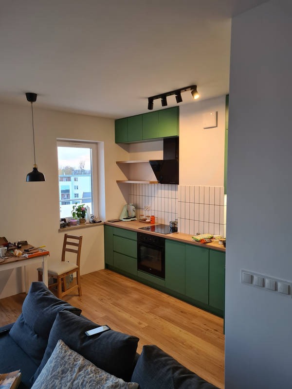 Wnętrze mieszkania z otwartą kuchnią: zielone szafki kuchenne z drewnianym blatem, białe płytki na ścianie, drewniana podłoga, widok z salonu na okno i stół roboczy.