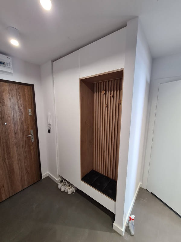 Biała szafa wnękowa z drewnianymi lamelami, siedziskiem i ukrytymi butami w przedpokoju.