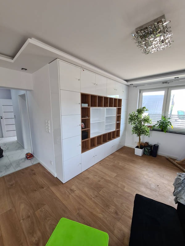 Biała zabudowa meblowa na wymiar z otwartymi półkami z drewna, w tle okno z roślinami i nowoczesny żyrandol.