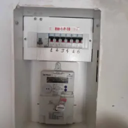 Zamknięta skrzynka rozdzielcza z wyłącznikami i licznikiem energii elektrycznej zamontowana w ścianie wnęki, z ręcznie dopisanymi numerami nad wyłącznikami.
