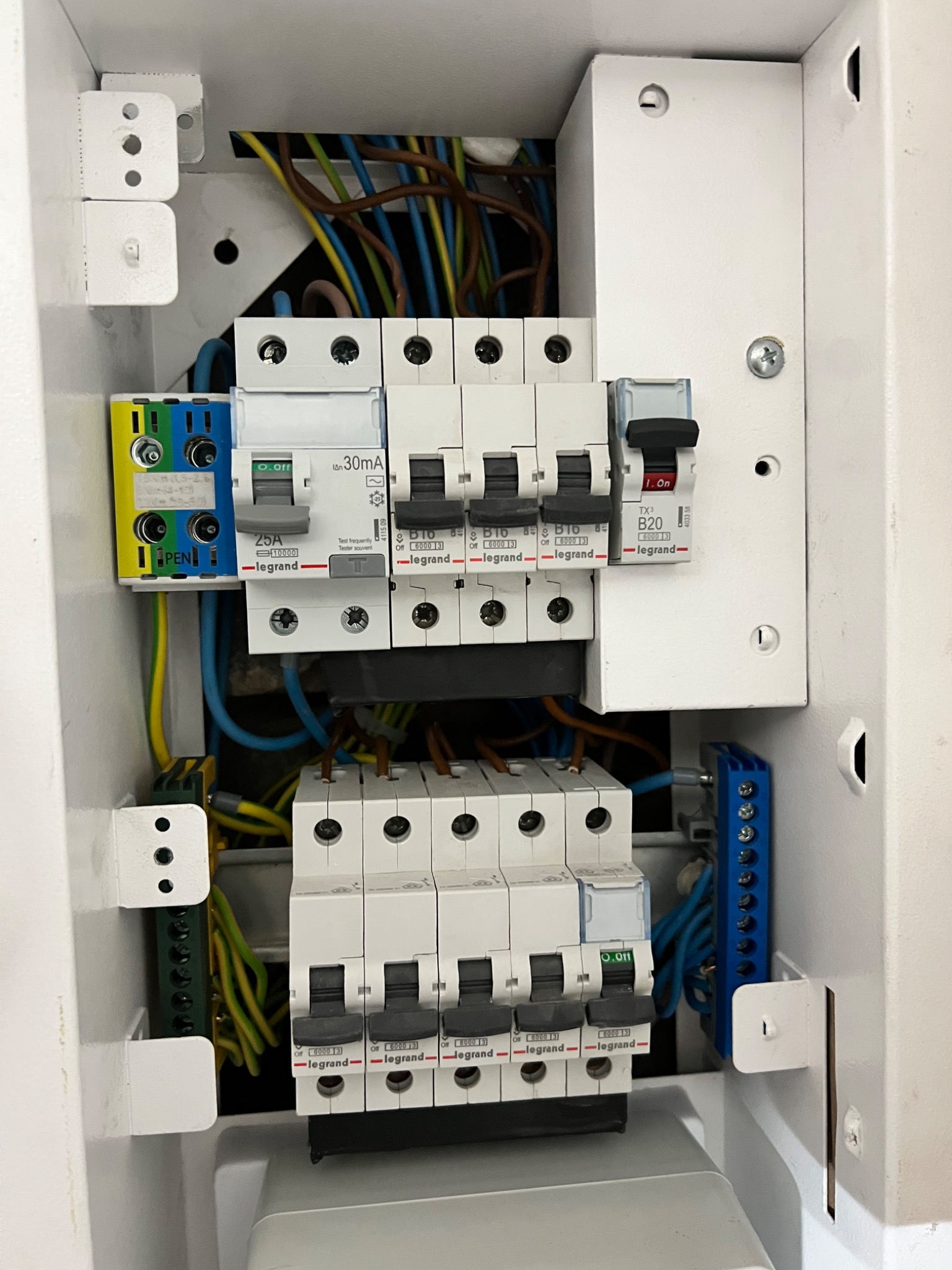 Wnętrze białej skrzynki rozdzielczej z zainstalowanymi bezpiecznikami i przewodami elektrycznymi różnych kolorów, widoczne oznaczenia Legrand na elementach.