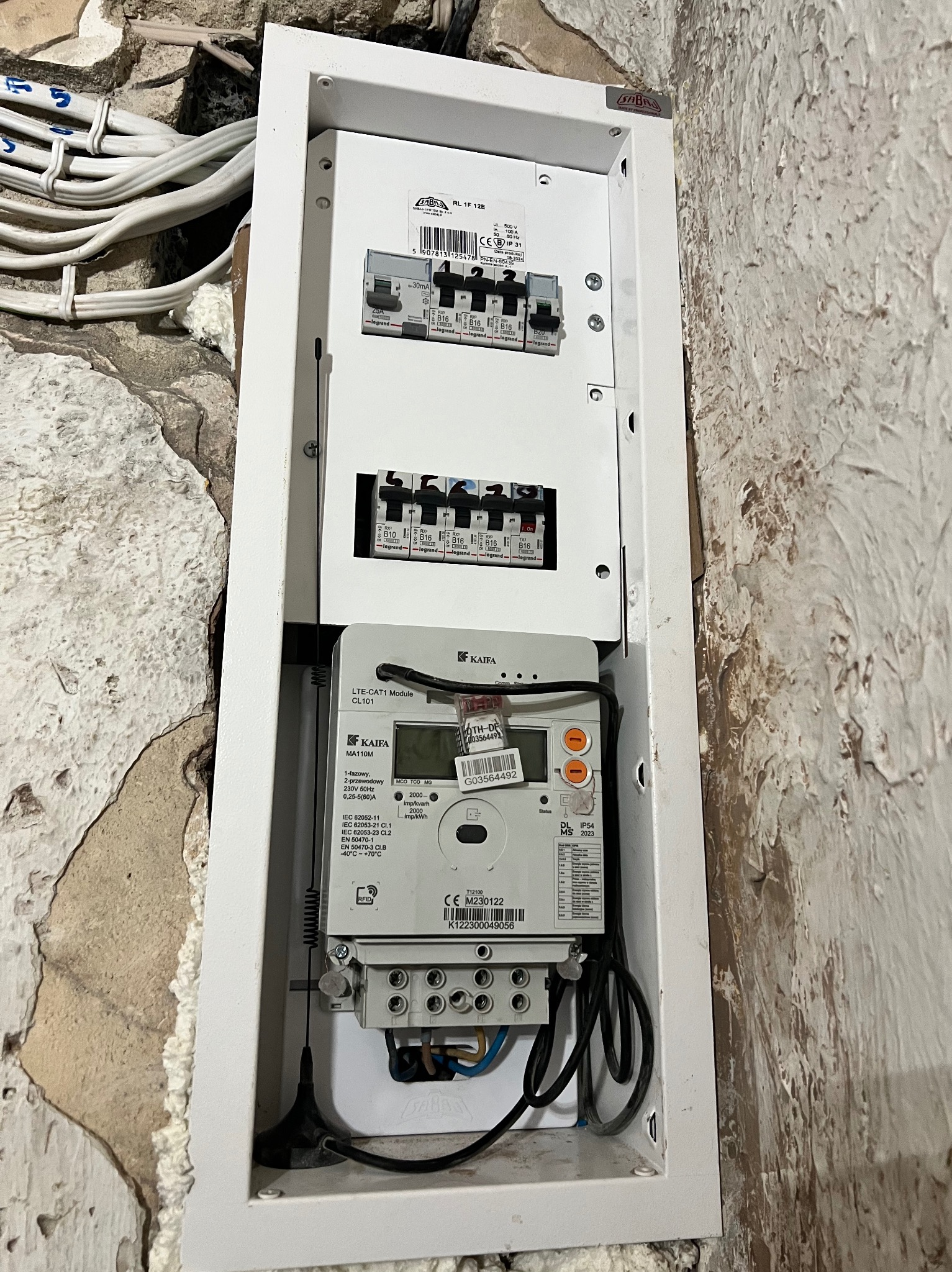 Otwarta skrzynka elektryczna z zamontowanym licznikiem energii i zabezpieczeniami, widoczne przewody i fragment ściany z surowym wykończeniem.