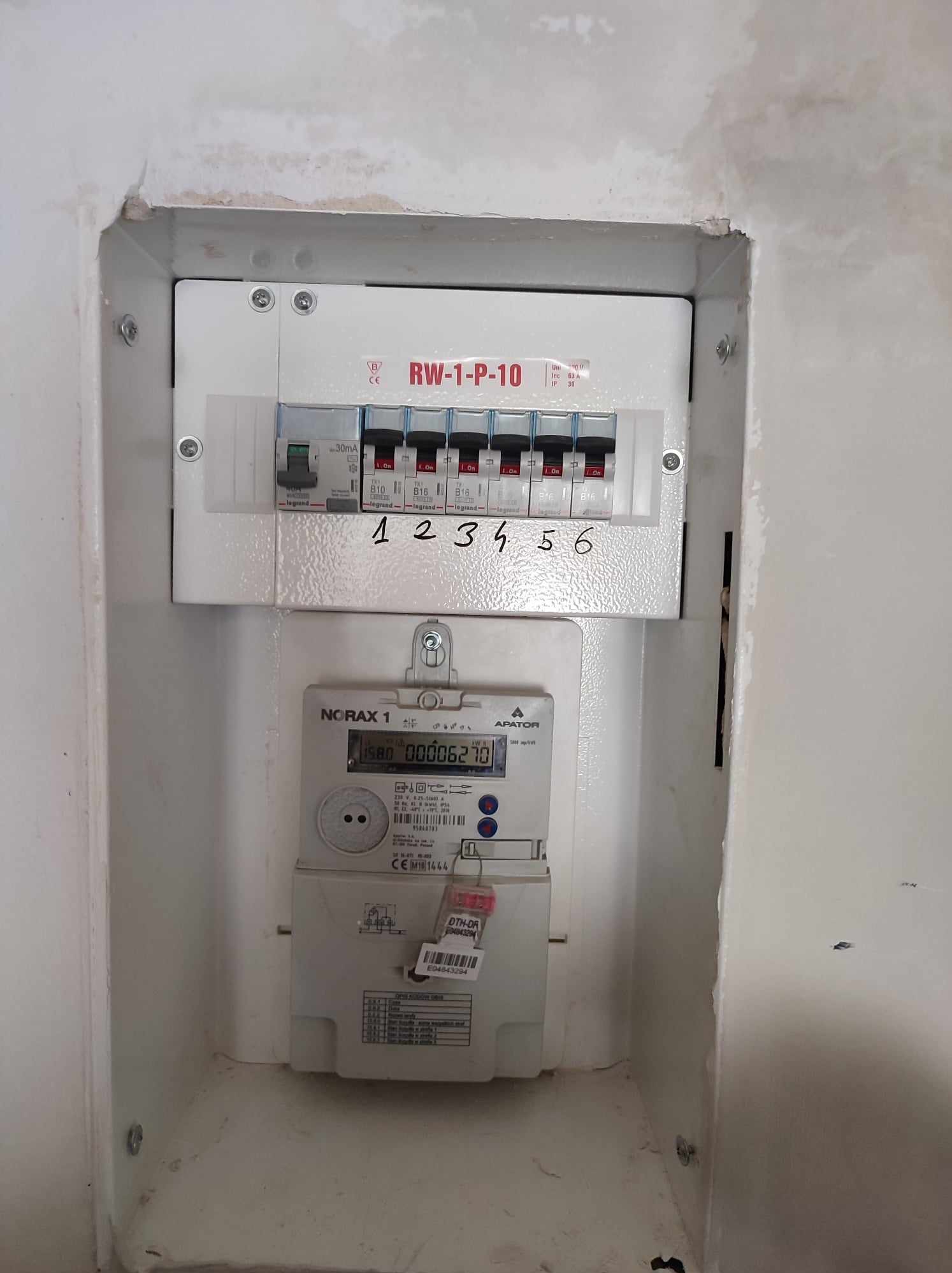 Zamknięta skrzynka rozdzielcza z wyłącznikami i licznikiem energii elektrycznej zamontowana w ścianie wnęki, z ręcznie dopisanymi numerami nad wyłącznikami.