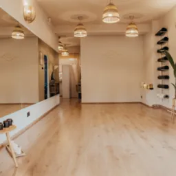 Jasne, przestronne wnętrze studia jogi z drewnianą podłogą, dużym lustrem, dekoracyjnymi lampami z rattanu, ścianami w odcieniach beżu i minimalistycznymi dekoracjami.