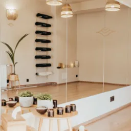 Jasne wnętrze studia jogi z dużym lustrem, drewnianą podłogą, rattanowymi lampami i półką na maty do ćwiczeń.