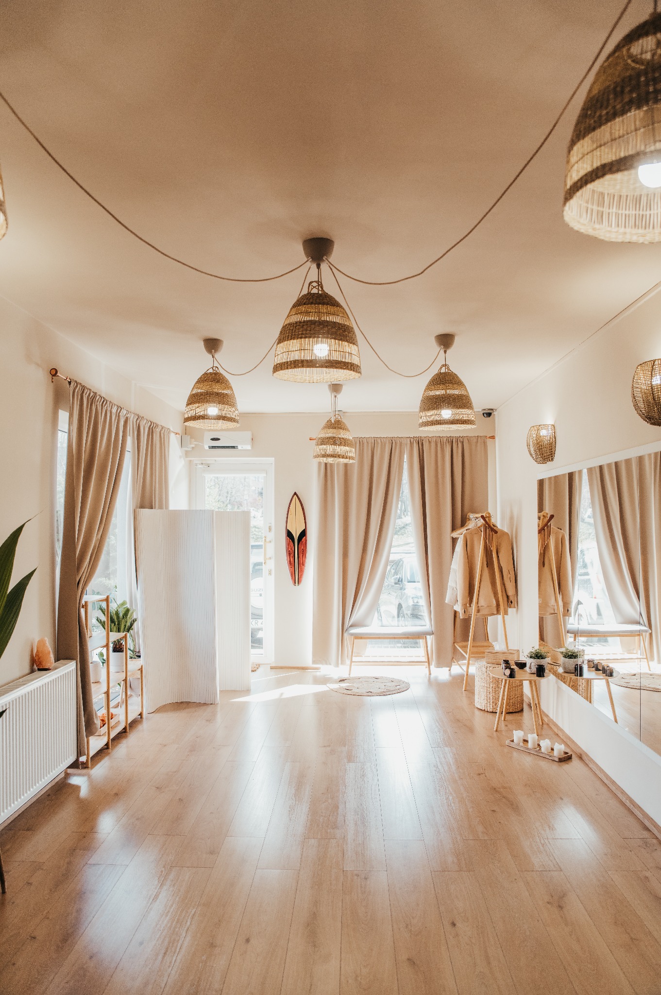 Jasne wnętrze w stylu boho z drewnianą podłogą, wiklinowymi lampami, parawanem, lustrem i dekoracjami w postaci deski surfingowej i roślin.