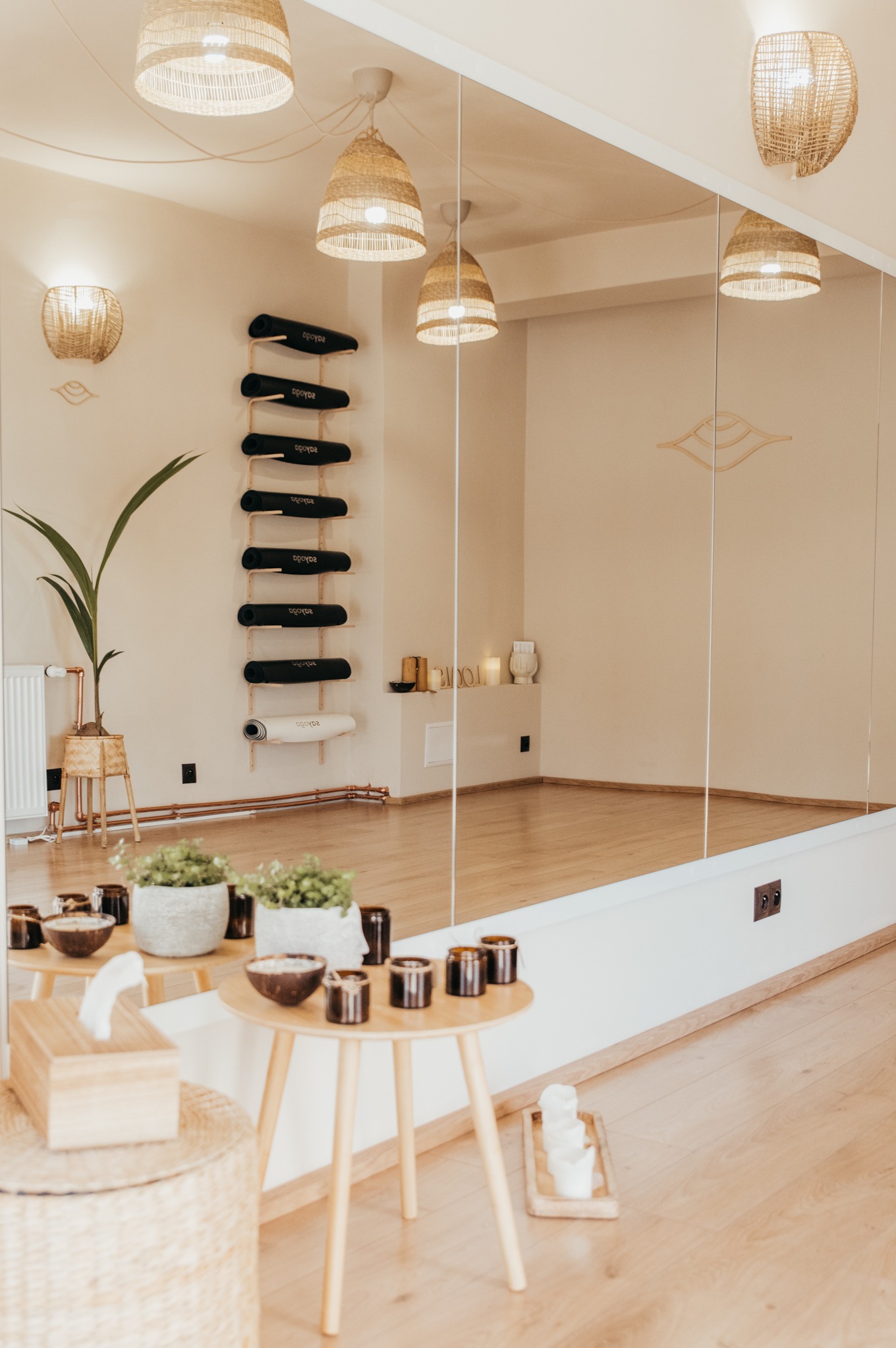 Jasne wnętrze studia jogi z dużym lustrem, drewnianą podłogą, rattanowymi lampami i półką na maty do ćwiczeń.