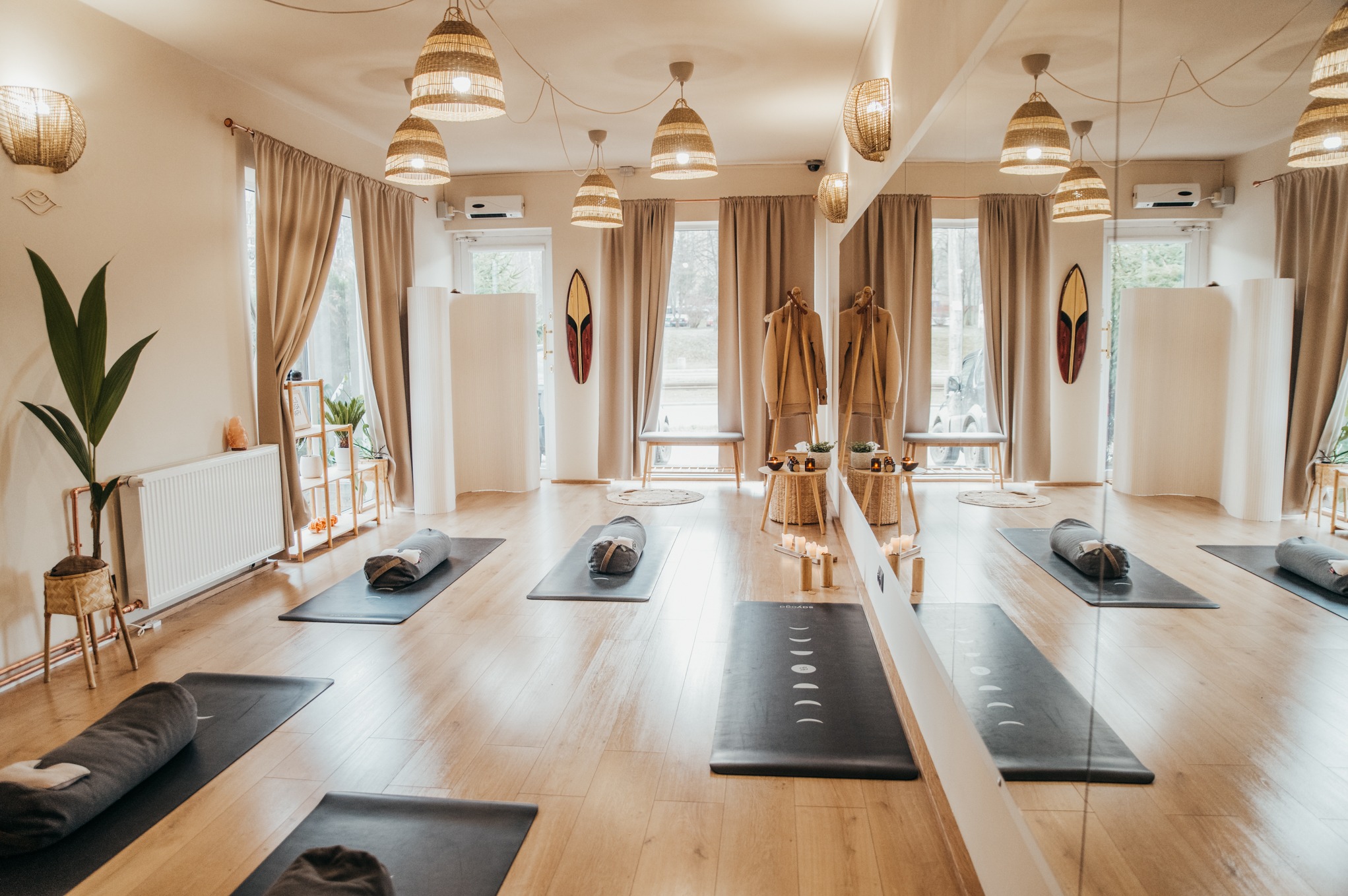 Jasne, przestronne studio jogi z drewnianą podłogą, matami do ćwiczeń, dekoracyjnymi lampami z wikliny, dużym lustrem i roślinami doniczkowymi, tworzące spokojną atmosferę.