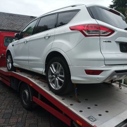 Usługi transportowe Taxi - Biały samochód Ford Kuga na czerwonej lawecie z widocznymi hakami zabezpieczającymi koła podczas transportu, w tle budynek z szarą dachówką.