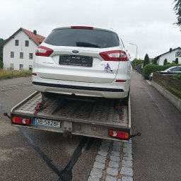 Usługi transportowe Taxi - Biały Ford Mondeo na lawecie w Wałbrzychu. Na tylnej szybie widoczna naklejka z wizerunkiem wiedźmy. Laweta zaparkowana na wąskiej ulicy, w tle zabudowa jednorodzinna.