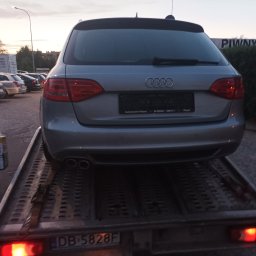 Usługi transportowe Taxi - Srebrne Audi kombi, widok z tyłu, załadowane na lawetę z włączonymi światłami pozycyjnymi, mężczyzna w tle trzyma pudełko, na parkingu o zmierzchu.