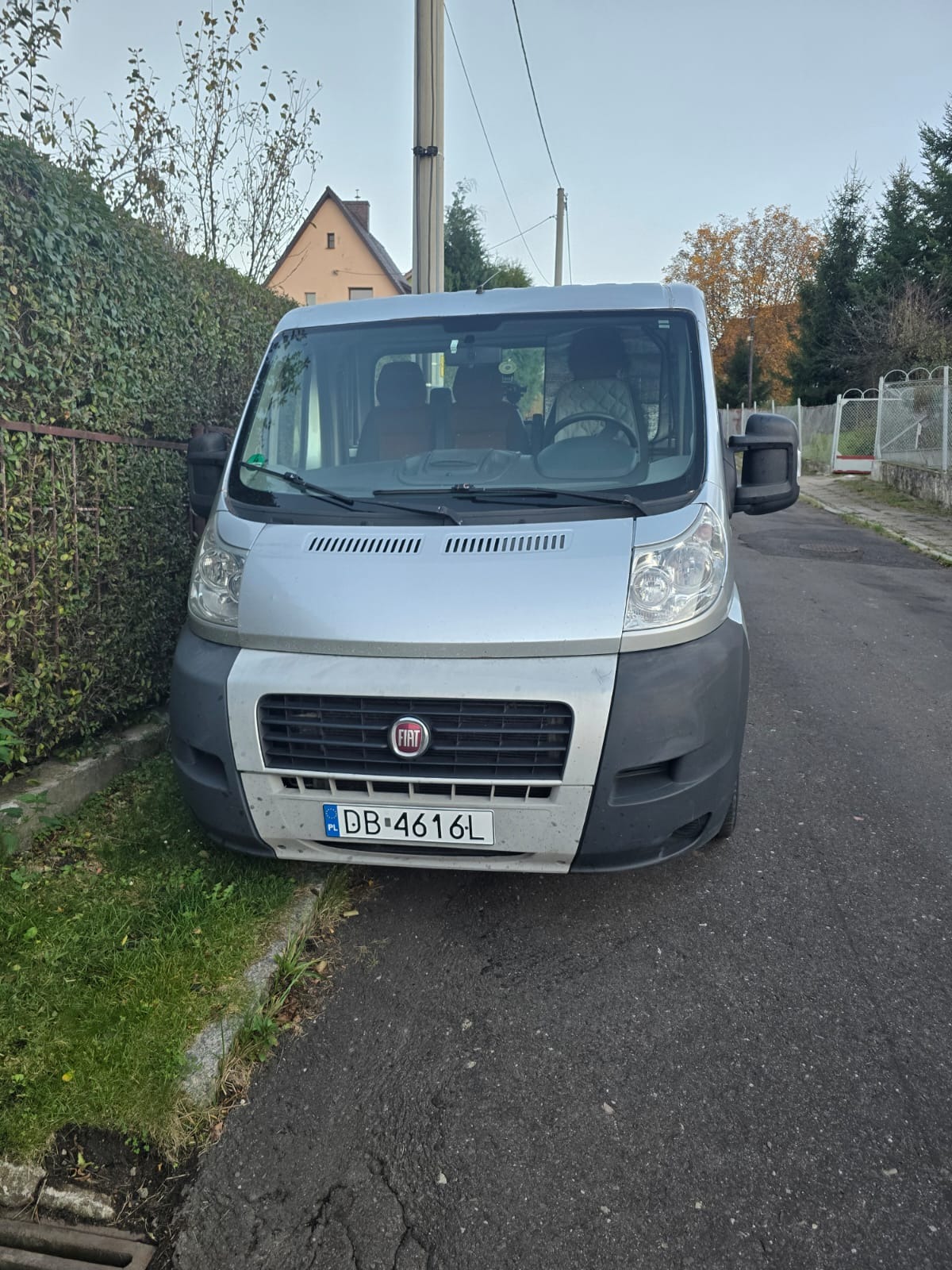 Srebrny bus marki Fiat Ducato zaparkowany na ulicy, widok z przodu, z widoczną tablicą rejestracyjną DB 4616L, obok zielony żywopłot i słup elektryczny.