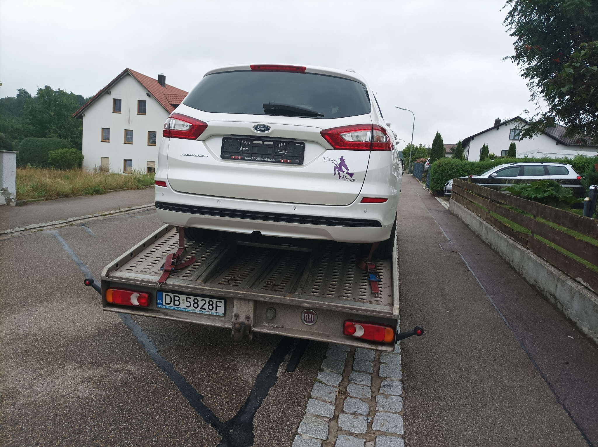 Biały Ford Mondeo na lawecie w Wałbrzychu. Na tylnej szybie widoczna naklejka z wizerunkiem wiedźmy. Laweta zaparkowana na wąskiej ulicy, w tle zabudowa jednorodzinna.