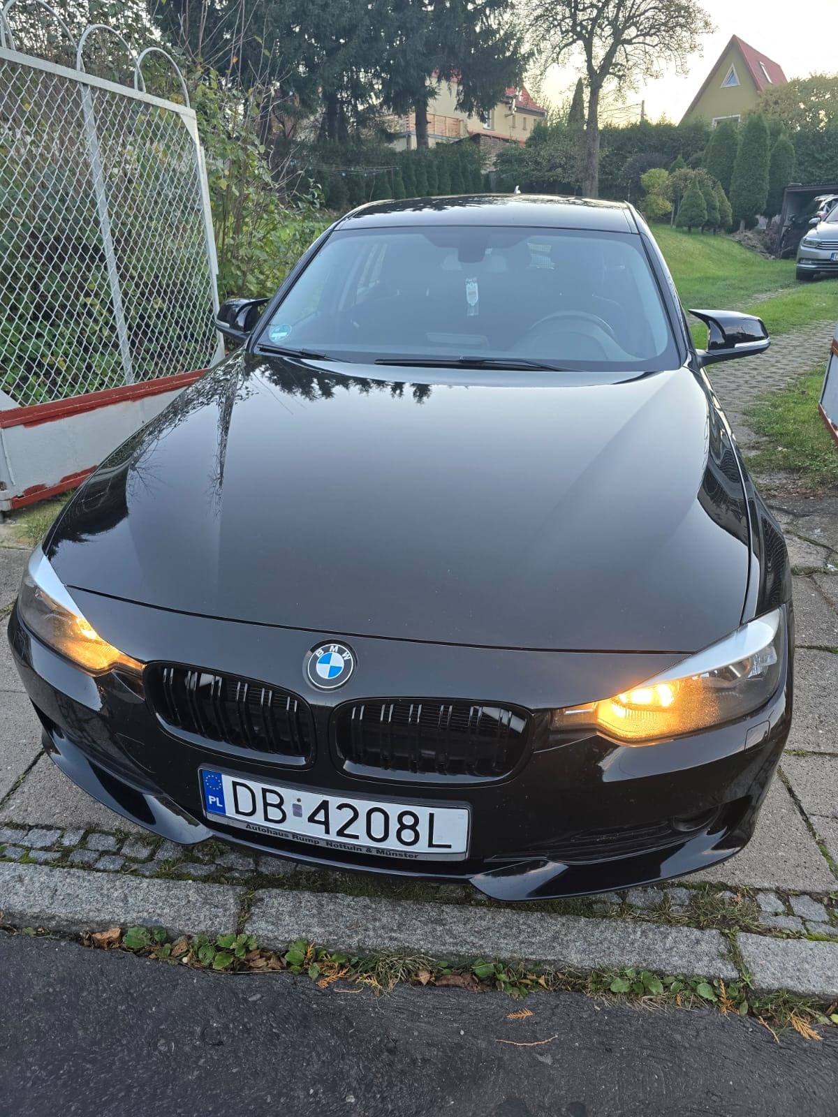 Czarna limuzyna BMW serii 3, zaparkowana na brukowanym podjeździe przed domem z zielonym trawnikiem i ogrodzeniem z siatki, z włączonymi światłami mijania i widoczną polską tablicą rejestracyjną.