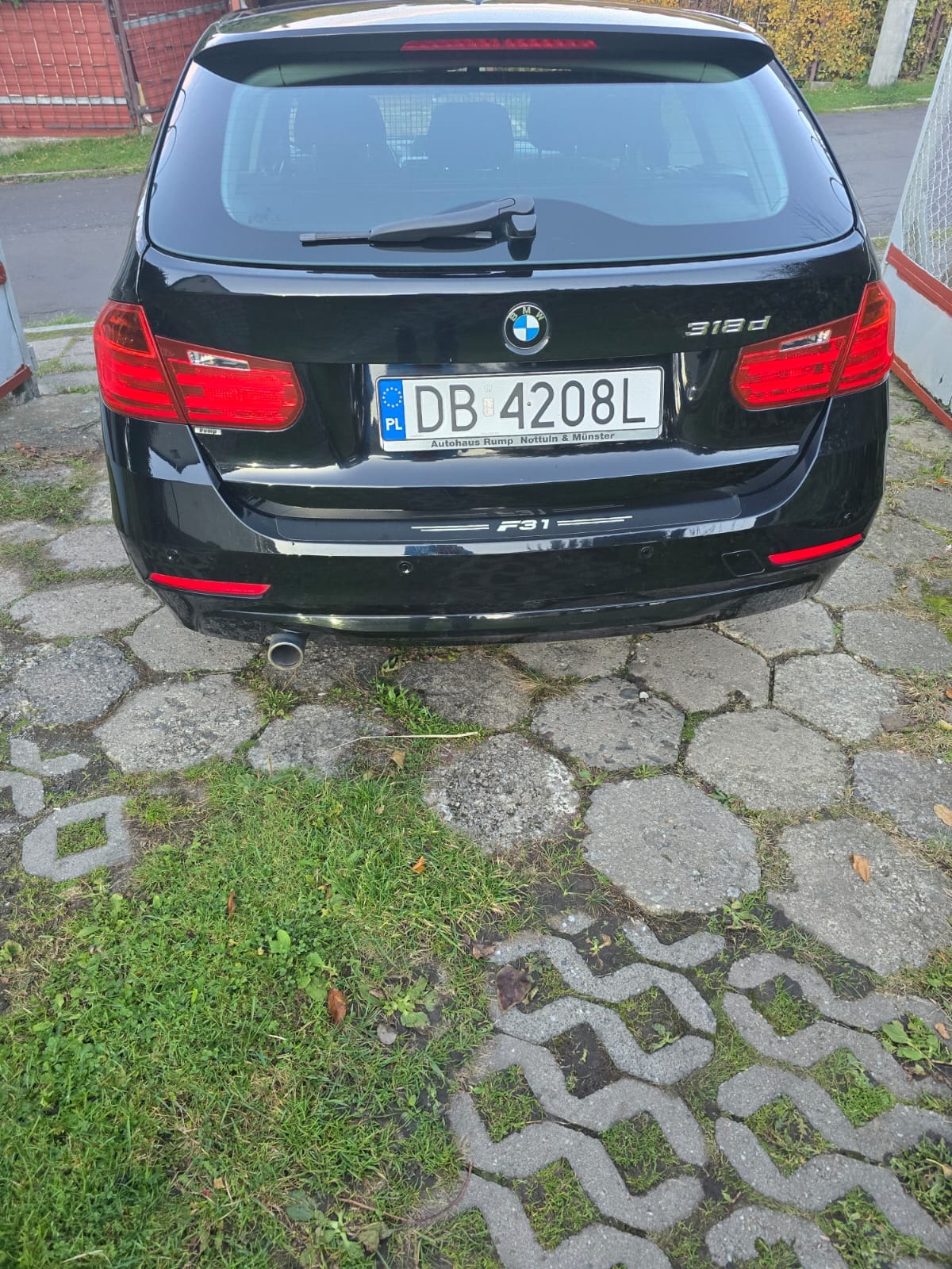 Czarny samochód BMW 318d kombi, widok z tyłu, tablica rejestracyjna DB 4208L, zaparkowany na kostce brukowej z fragmentami trawy.