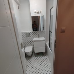 Stan - Wąskie ujęcie wyremontowanej łazienki z toaletą, umywalką z szafką i kabiną prysznicową, wykończonej białymi płytkami z czarnym wzorem na podłodze i części ściany, lustro odbija osobę robiącą zdjęcie.