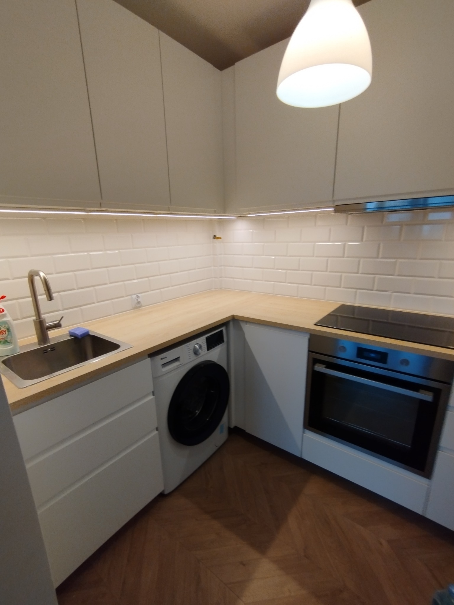 Nowoczesna, narożna kuchnia z białymi szafkami, blatem z jasnego drewna, białymi płytkami na ścianie, zintegrowaną pralką i piekarnikiem, oraz parkietem ułożonym w jodełkę na podłodze.