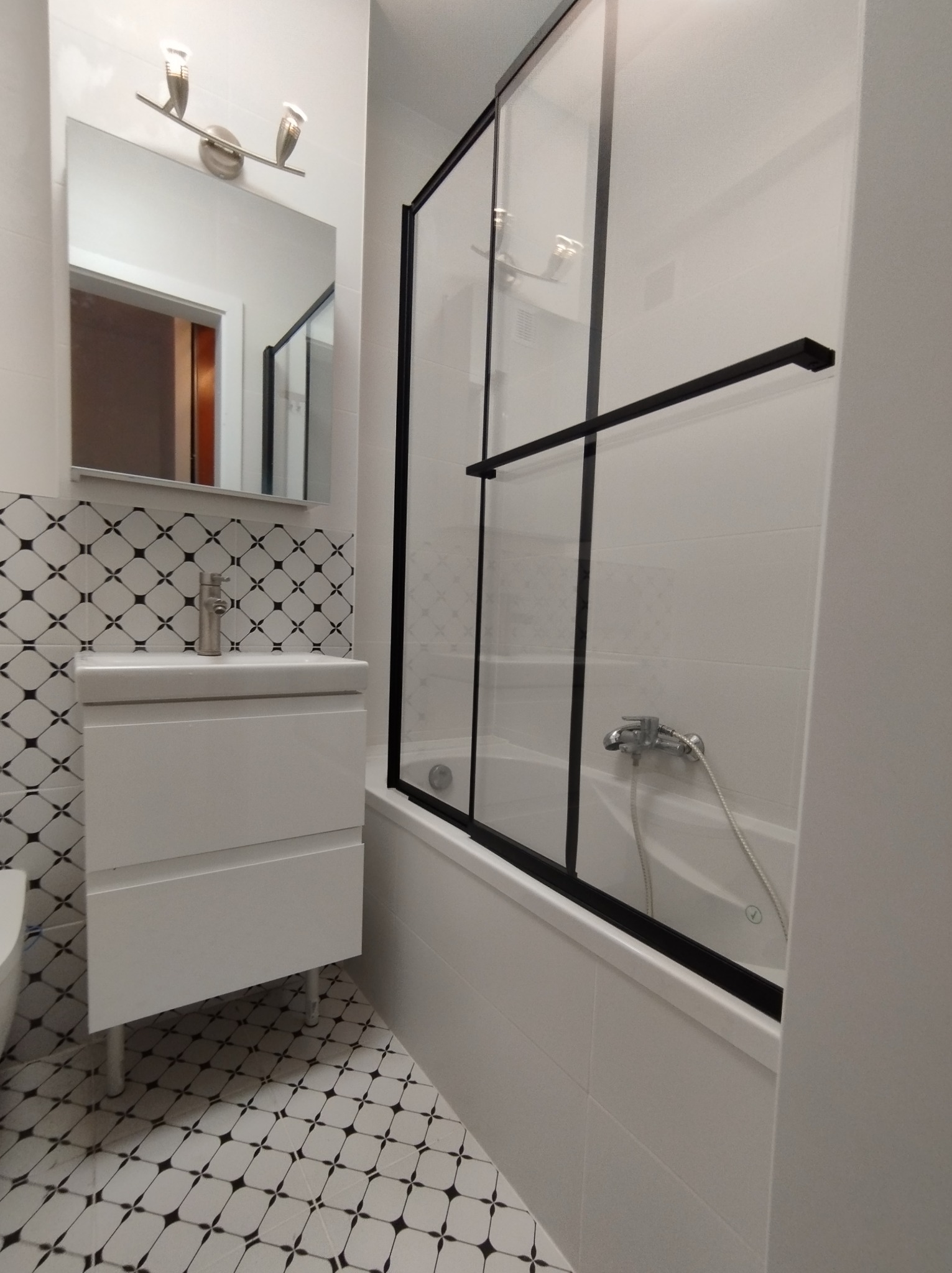 Nowoczesna łazienka w bieli i czerni: umywalka z szafką na nóżkach, bateria i lustro na tle mozaiki, kabina prysznicowa z czarnymi ramami, białe płytki na ścianach i podłodze z wzorem karo.