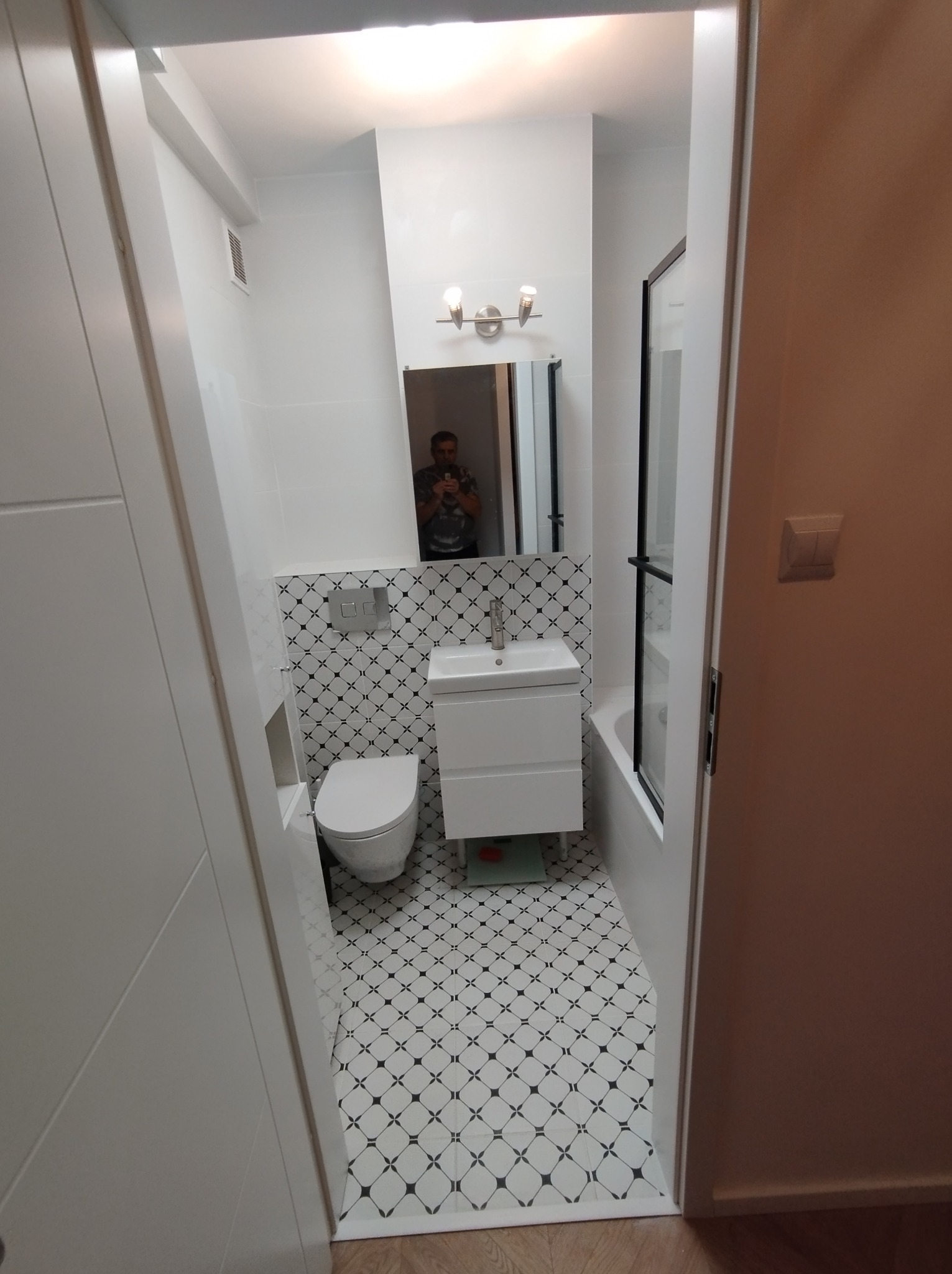 Wąskie ujęcie wyremontowanej łazienki z toaletą, umywalką z szafką i kabiną prysznicową, wykończonej białymi płytkami z czarnym wzorem na podłodze i części ściany, lustro odbija osobę robiącą zdjęcie.