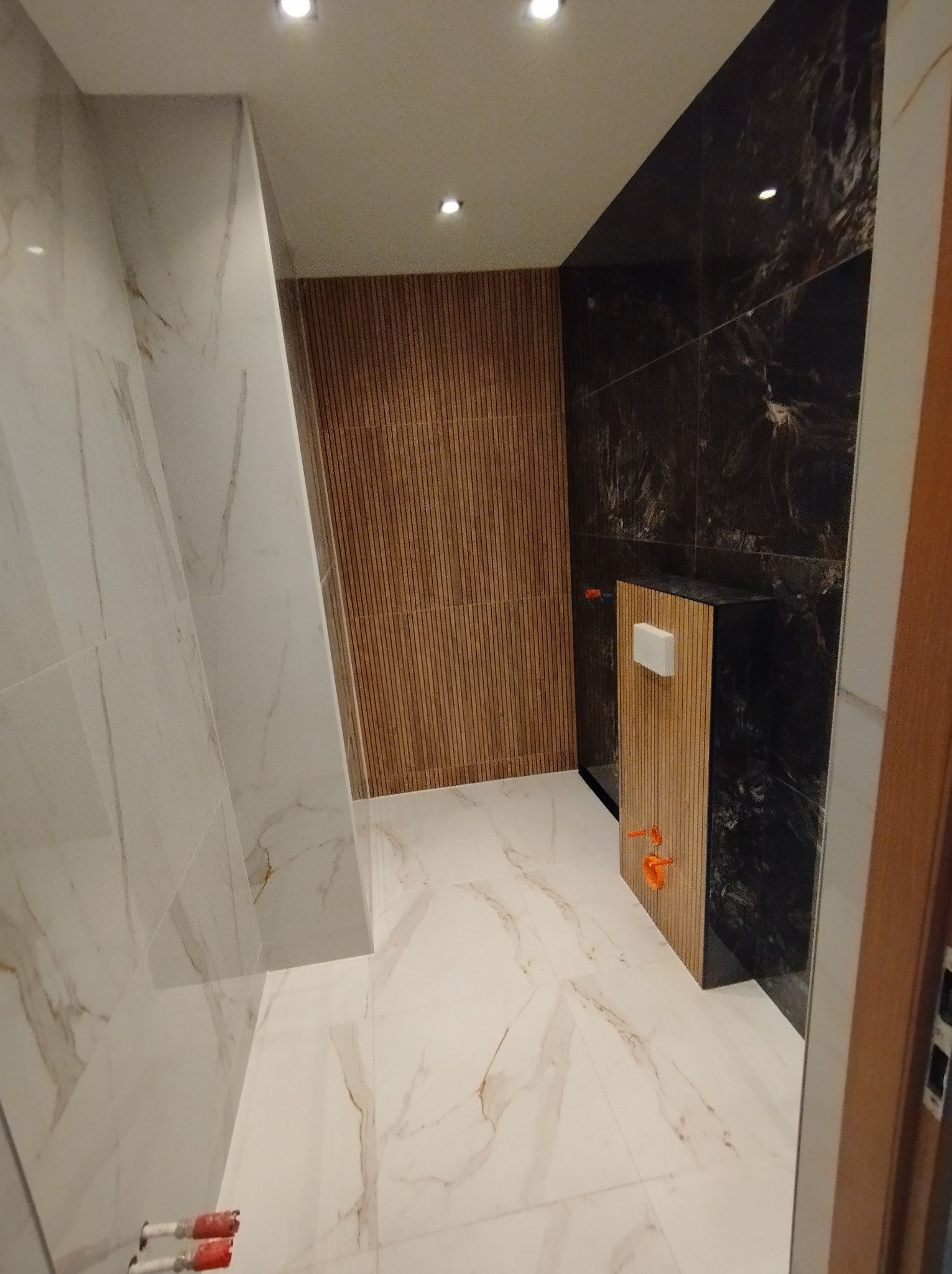 Nowoczesna łazienka z marmurowymi płytkami na ścianach i podłodze, drewnianą ścianą z pionowych listew oraz zabudowanym stelażem WC z elementami instalacji hydraulicznej.