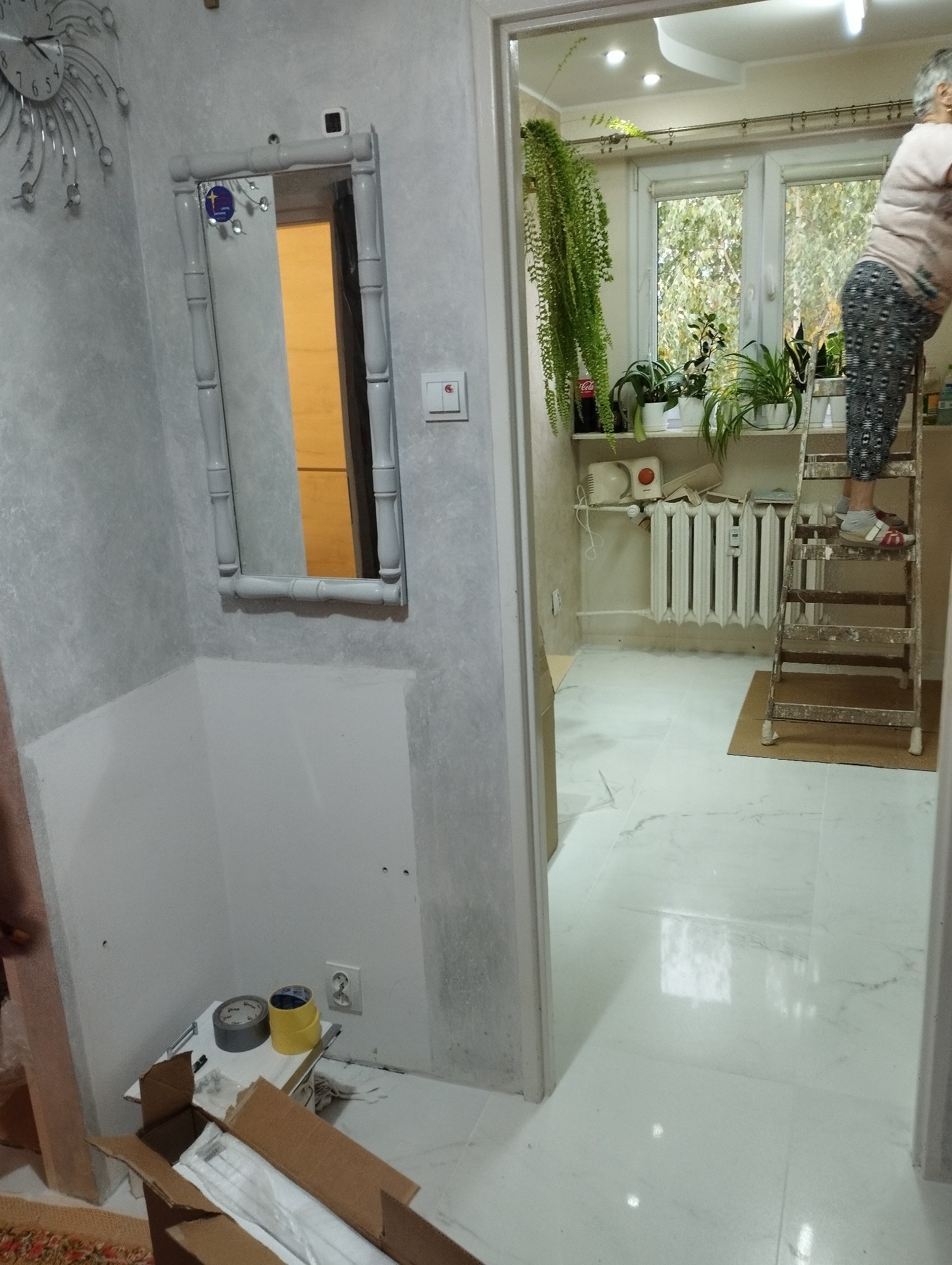 Korytarz w trakcie odświeżania, widoczna ściana z lustrem w bambusowej ramie, częściowo odmalowana, z widocznymi śladami po wcześniejszej aranżacji, w tle osoba na drabinie przy oknie z roślinami.