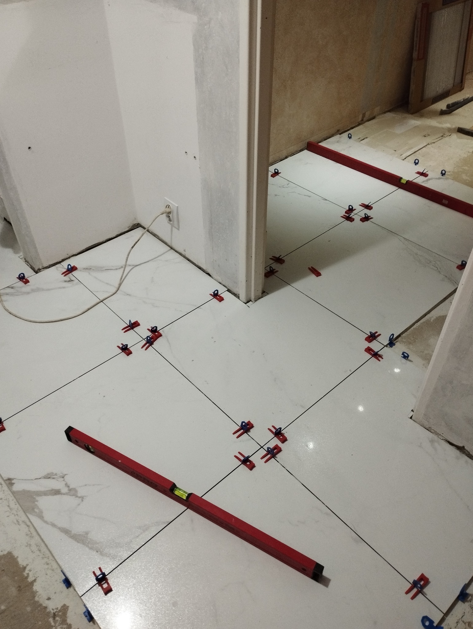 Białe płytki podłogowe w trakcie układania, z użyciem czerwonych i niebieskich klipsów poziomujących, widoczna poziomica, surowy stan ścian.