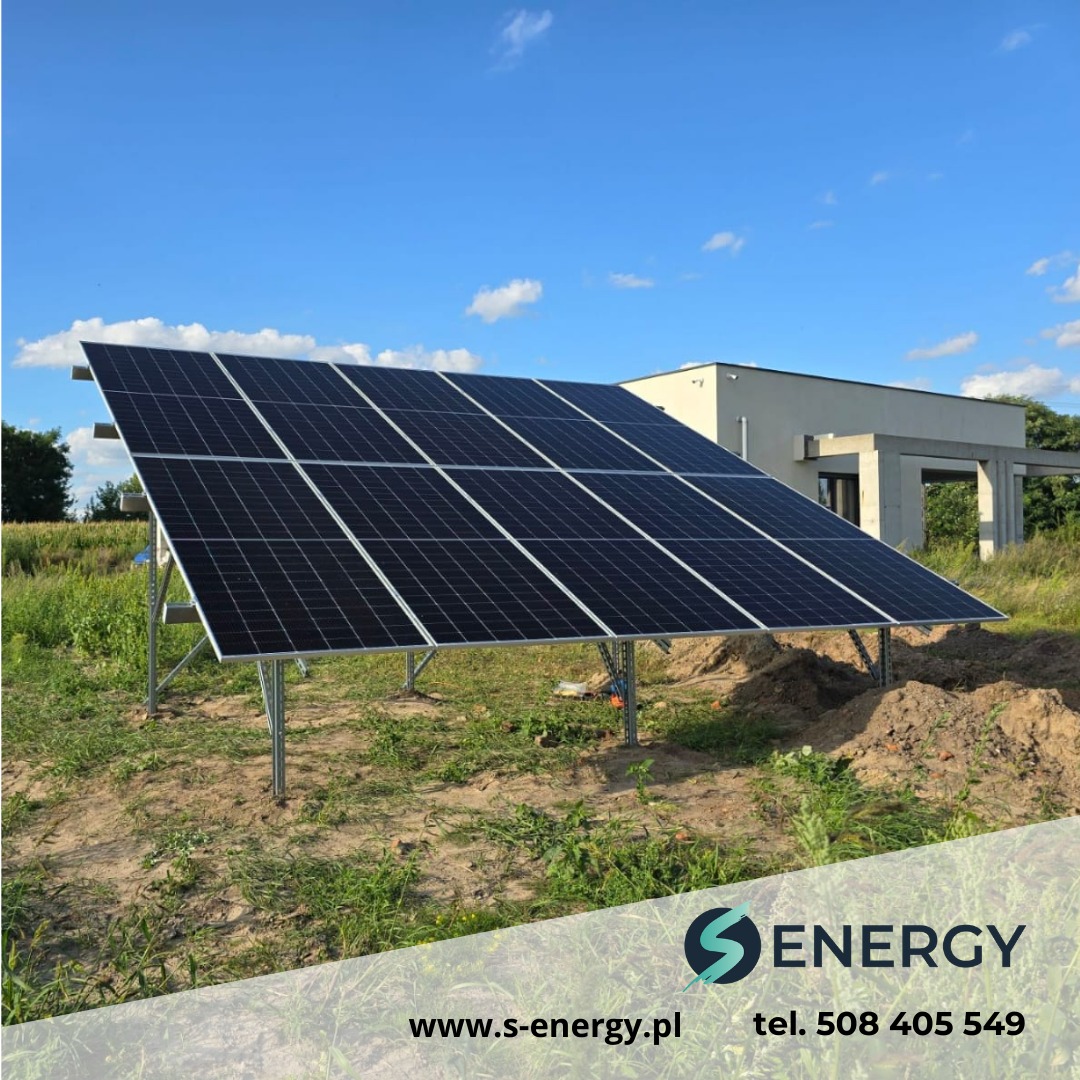 Instalacja paneli fotowoltaicznych na gruncie w Wołominie, widoczne metalowe konstrukcje wsporcze i budynek w tle pod błękitnym niebem.