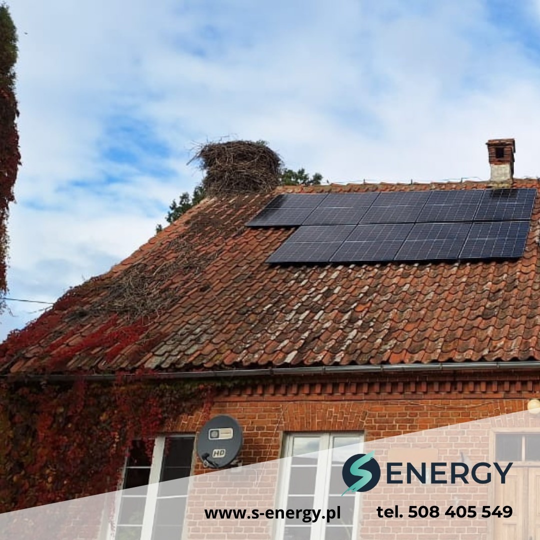Panele słoneczne na dachu starego, ceglanego domu z gniazdem bocianim, widziane pod kątem. Częściowo zasłonięte logo firmy z danymi kontaktowymi.