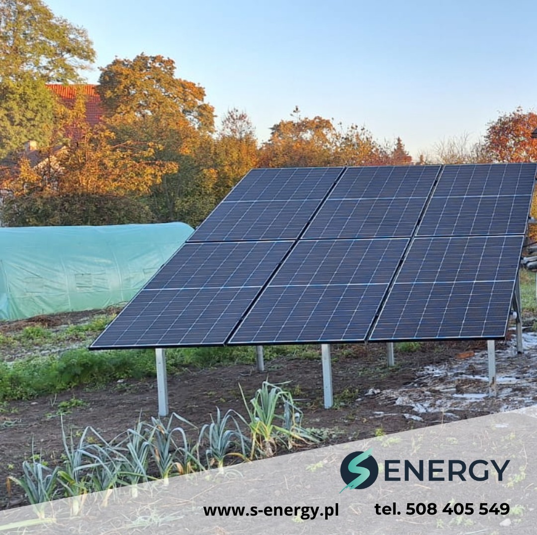 Instalacja paneli słonecznych na gruncie w Wołominie. Widoczne sześć paneli, konstrukcja wsporcza, ogród warzywny w tle i fragment tunelu foliowego.