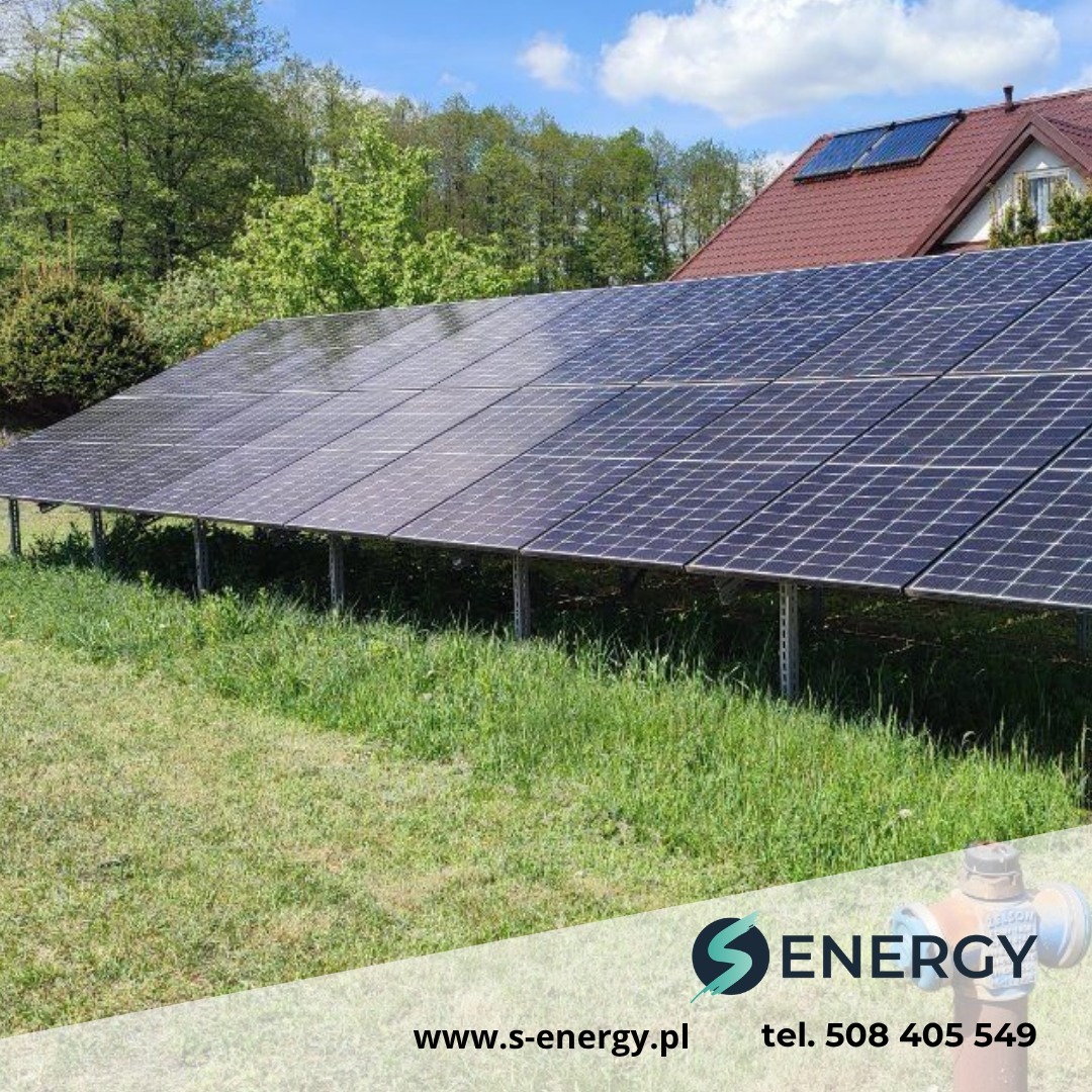 Instalacja fotowoltaiczna na trawiastym terenie, panele skierowane w stronę nieba. Na dachu domu widoczny dodatkowy panel solarny.