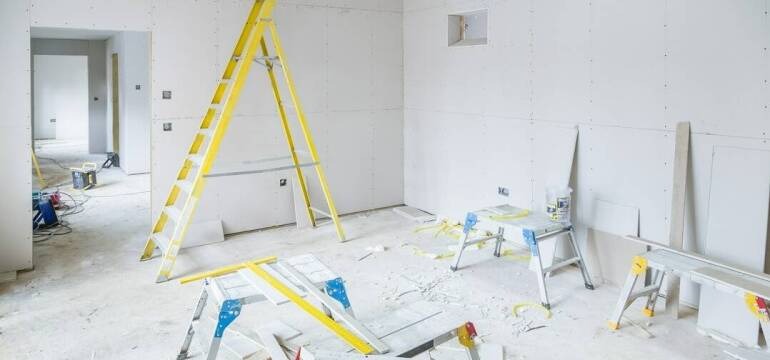 Pokój w trakcie montażu płyt gipsowo-kartonowych, widoczne żółte drabiny, białe płyty, szpachla i narzędzia na podłodze.