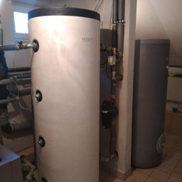 BC Karol Żuradzki - Biały, pionowy zbiornik akumulacyjny Netsu z czarną górą, obok szary zbiornik, rury w izolacji, na tle beżowych płytek podłogowych i białych ścian.