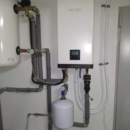 BC Karol Żuradzki - Biały, wiszący magazyn energii Netsu z podłączonymi rurami i zbiornikiem wyrównawczym, zamontowany na jasnej ścianie w pomieszczeniu technicznym.