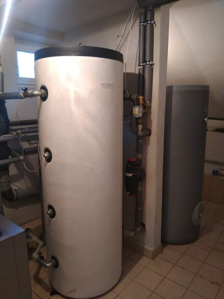 Biały, pionowy zbiornik akumulacyjny Netsu z czarną górą, obok szary zbiornik, rury w izolacji, na tle beżowych płytek podłogowych i białych ścian.